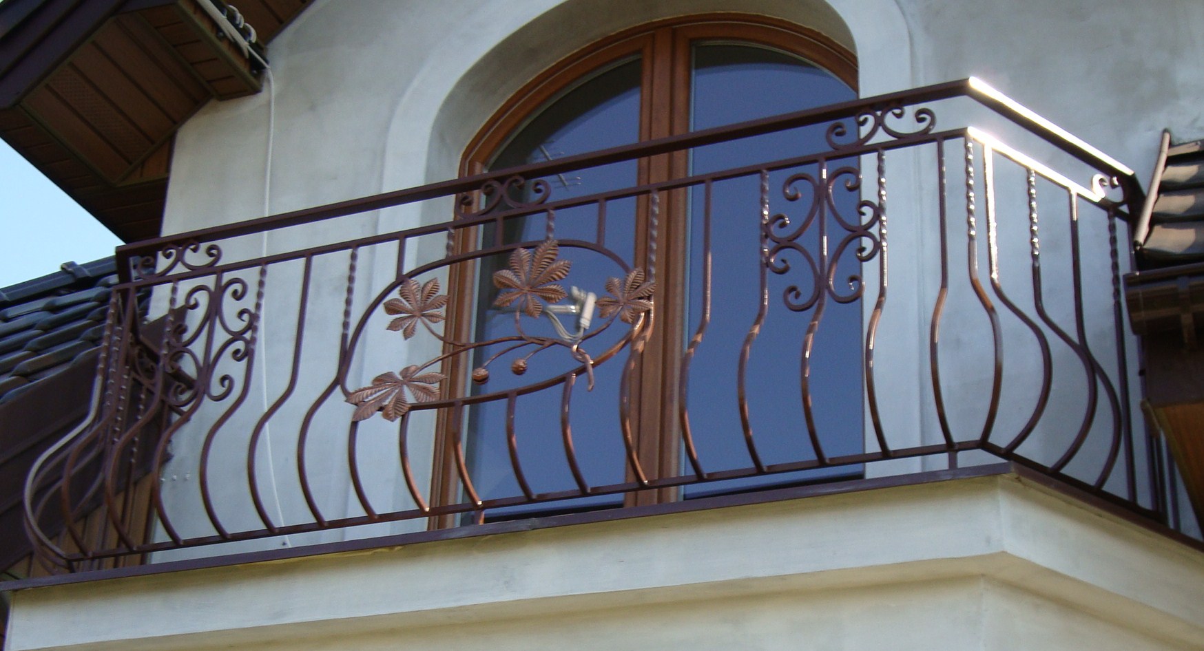 Kuta balustrada balkonowa z motywem liści kasztanowca, brązowa, na tle okna z łukiem i fragmentu dachu.